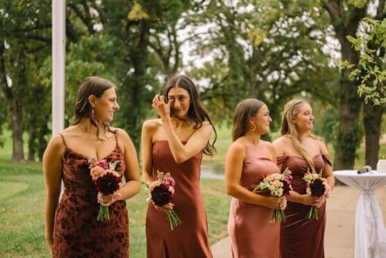 Four bridesmaids in matching rust-colored dresses stand outdoors, each holding a bouquet of flowers. One woman wipes away a tear while the others smile. Green trees and grass are visible in the background. by Justin Salem Meyer