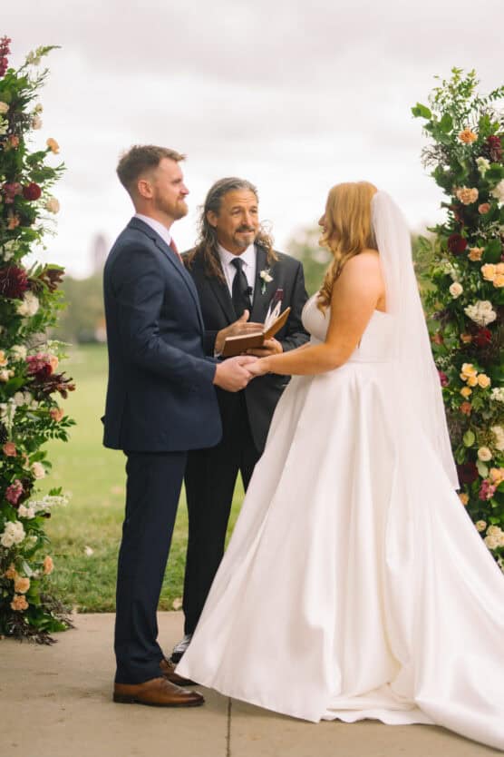 A bride and groom stand facing each other, holding hands and smiling, with a wedding officiant between them. They are outdoors, framed by floral arches, with greenery in the background. by Justin Salem Meyer