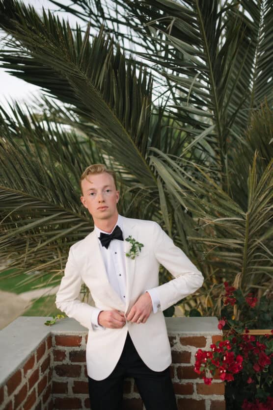 A young man in a white tuxedo jacket and black bow tie stands in front of palm leaves and a brick wall with red flowers, looking slightly to the side with a calm expression. by Justin Salem Meyer