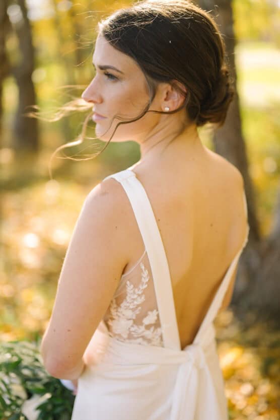 A woman in a sleeveless wedding dress with lace details and an open back stands outdoors, looking over her shoulder. Her dark hair is styled in a low updo, and sunlight filters through trees in the background. by Justin Salem Meyer