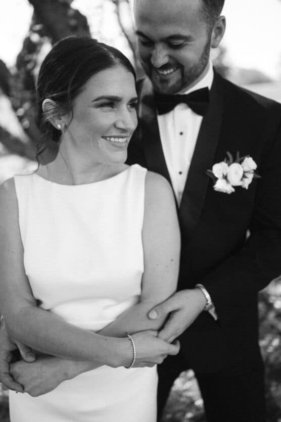 A smiling bride in a sleeveless white dress stands in front of a groom in a tuxedo, who embraces her from behind. They appear happy and affectionate outdoors. The image is in black and white. by Justin Salem Meyer