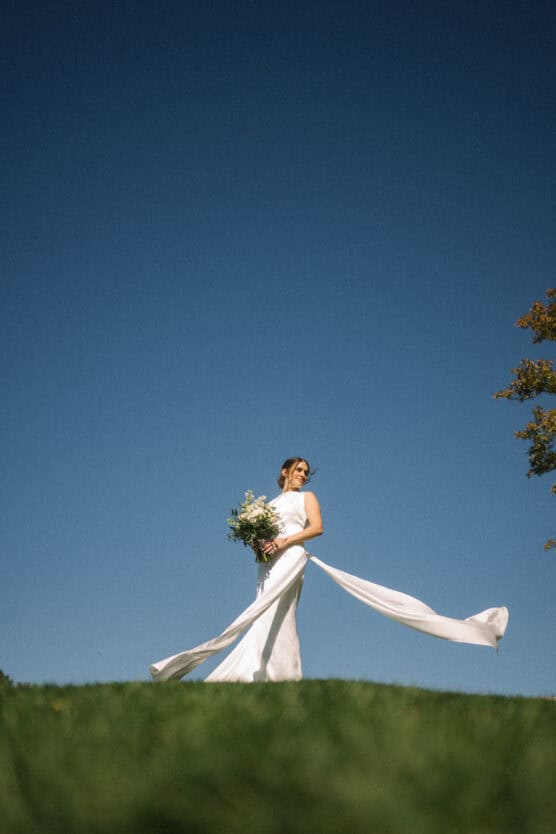 A bride in a flowing white dress stands on green grass, holding a bouquet of flowers against a clear blue sky. Her dress billows in the breeze, and sunlight illuminates the scene. by Justin Salem Meyer