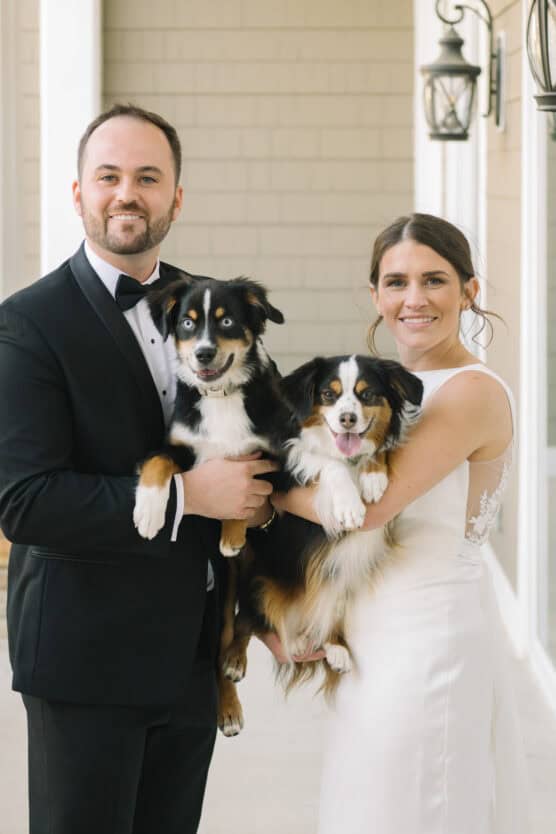 A bride in a white dress and a groom in a black tuxedo stand side by side, each holding a happy black, brown, and white dog, posing together and smiling in front of a building with beige siding and lantern lights. by Justin Salem Meyer