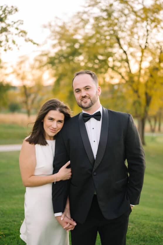 A woman in a white sleeveless dress stands closely beside a man in a black tuxedo, holding hands. They are outdoors on a grassy area with trees and soft evening light in the background. by Justin Salem Meyer