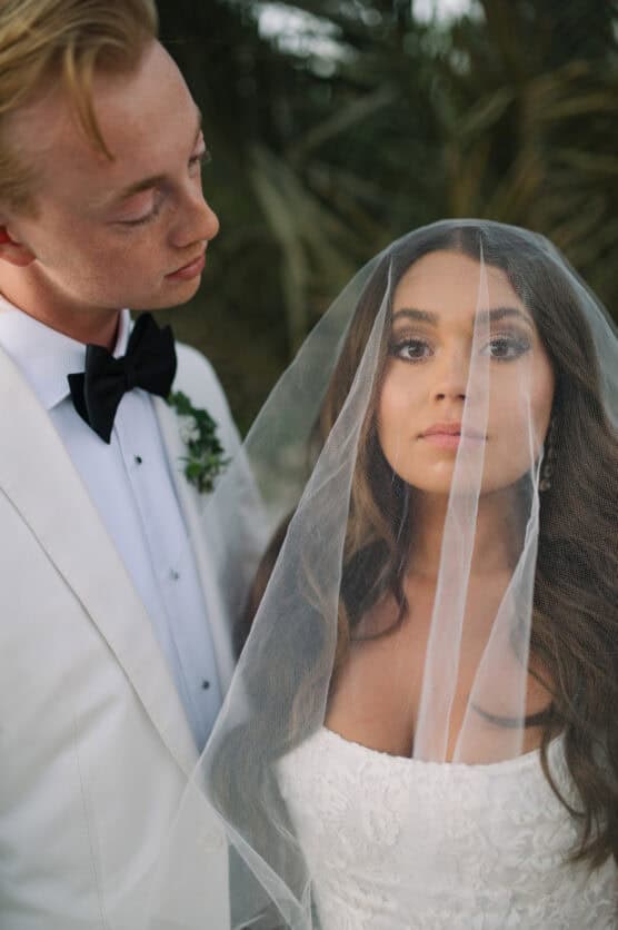A bride in a white dress and veil looks straight ahead while a groom in a white tuxedo jacket and black bow tie gazes at her lovingly. They are standing outdoors in front of palm leaves. by Justin Salem Meyer
