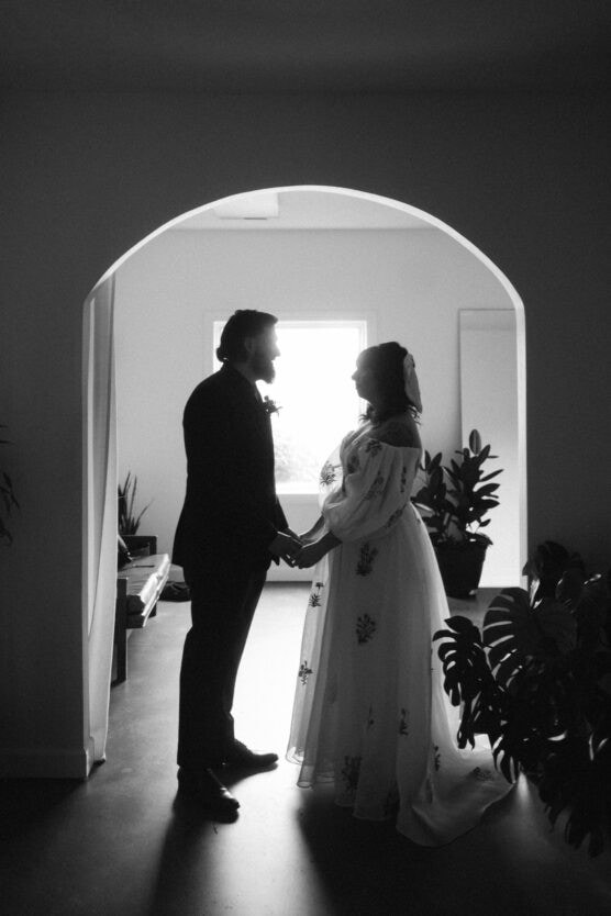 A black and white photo of a couple standing face-to-face under an arched doorway, holding hands. The woman wears a long dress and veil, and the man wears a suit. They are surrounded by potted plants with light behind them. by Justin Salem Meyer