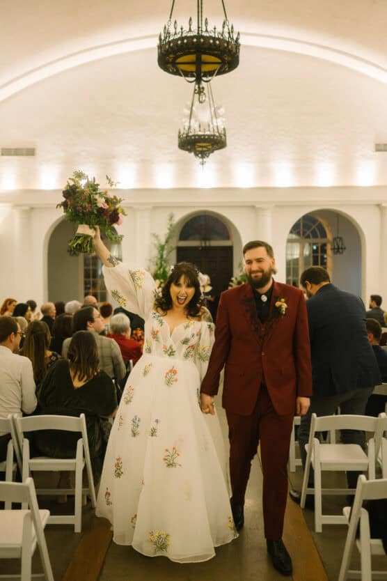 A joyful bride in a floral wedding dress holds up a bouquet while walking hand-in-hand with the groom in a burgundy suit down the aisle, surrounded by seated guests in a warmly lit indoor venue. by Justin Salem Meyer