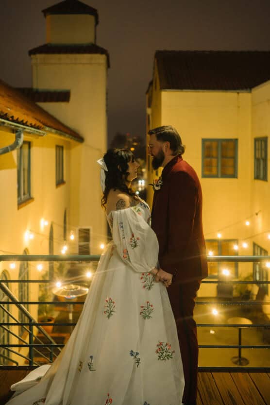 A bride and groom hold hands, facing each other on a balcony at night. The bride wears a white floral dress, and the groom wears a dark suit. Warm string lights and yellow buildings create a romantic, glowing backdrop. by Justin Salem Meyer