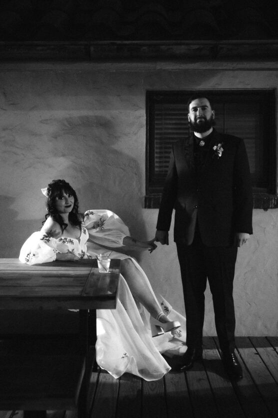 A bride in a flowing dress sits on a bench, reaching out to hold hands with a standing groom in a suit. The setting appears rustic with a stucco wall and a window behind them. The image is in black and white. by Justin Salem Meyer
