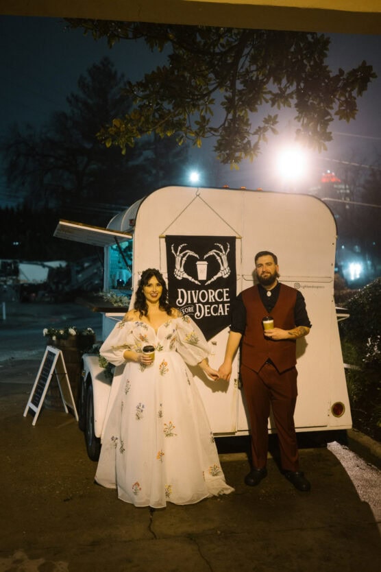 A couple stands holding hands in front of a white trailer with a sign that reads “Divorce for Decaf.” The woman wears a long white dress with floral patterns; the man wears a maroon suit and holds a drink. It appears to be nighttime. by Justin Salem Meyer