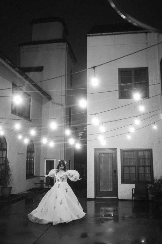 A person in a long, flowing dress stands under string lights in a courtyard at night, with tall white buildings in the background and reflections on the wet ground. The scene is in black and white. by Justin Salem Meyer