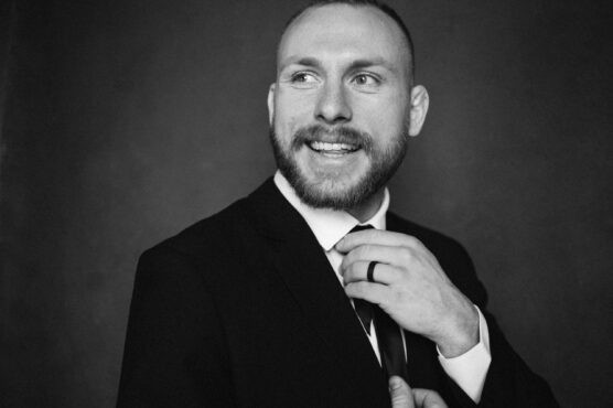 A man with a beard and short hair, dressed in a suit and tie, smiles while adjusting his tie. The photo is black and white with a dark, plain background. by Justin Salem Meyer