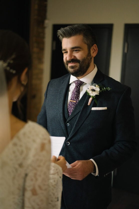 A groom in a dark suit with a floral boutonniere smiles warmly at his bride, who is holding a note. The bride is wearing a lace dress and veil, partially visible in the foreground. They are indoors, standing together. by Justin Salem Meyer