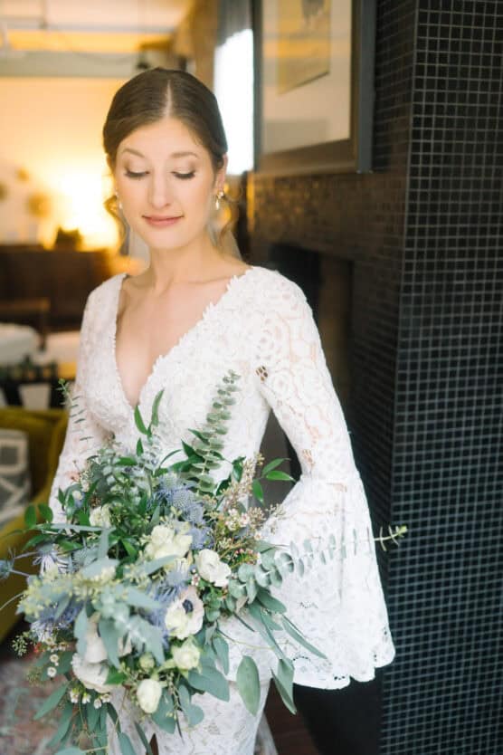 A bride in a long-sleeved white lace gown holding a large bouquet of greenery and white flowers, standing indoors with soft natural light and a calm expression. by Justin Salem Meyer