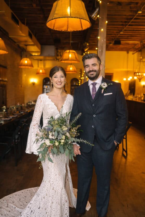 A bride in a lace gown holding a bouquet stands next to a groom in a dark suit and tie. They are smiling inside a warmly lit venue with wooden floors and hanging lamps. by Justin Salem Meyer