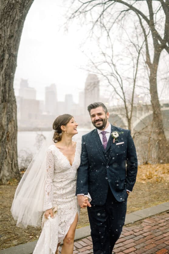 A bride in a lace dress and veil smiles at her groom, who wears a navy suit and tie. They walk hand in hand on a brick path outdoors with bare trees and city skyline in the snowy background. by Justin Salem Meyer