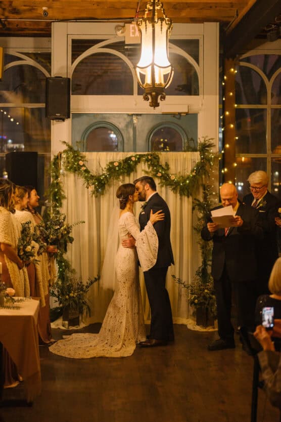A bride and groom kiss at their indoor wedding ceremony, surrounded by bridesmaids in light dresses, greenery decor, and officiants reading from papers under warm, romantic lighting. by Justin Salem Meyer