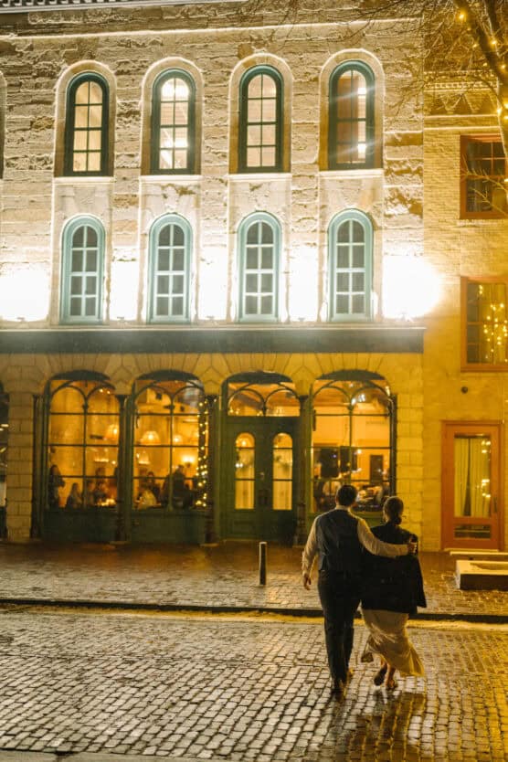 A couple walks arm in arm on a wet cobblestone street at night, passing a warmly lit building with tall arched windows, their backs turned to the camera. by Justin Salem Meyer