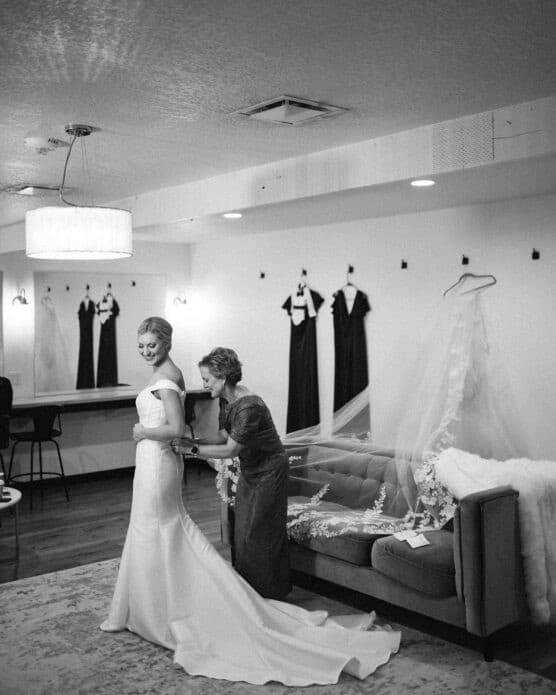 A bride smiles as someone helps her with her dress in a dressing room. Three dark dresses hang on the wall behind them, and a veil is draped over a sofa. The scene is elegant and softly lit. by Justin Salem Meyer