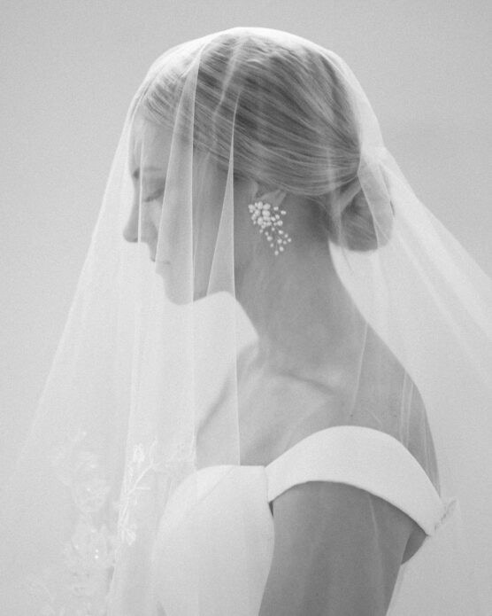 A black and white photo of a bride in profile, wearing a wedding dress with off-the-shoulder straps, a sheer veil over her face and hair, and ornate drop earrings. The image has a soft, dreamy quality. by Justin Salem Meyer