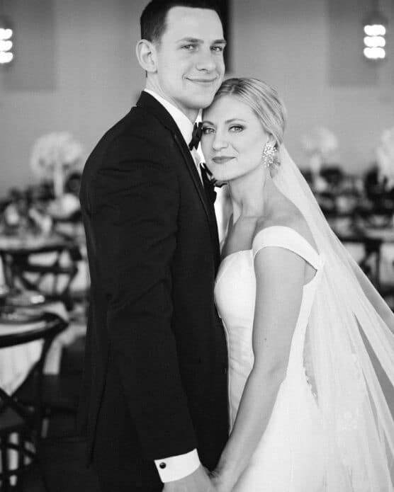 A bride and groom stand close together, holding hands and facing the camera. The groom wears a black tuxedo and the bride, in a white dress and veil, leans against him. The background shows a softly focused wedding venue. by Justin Salem Meyer