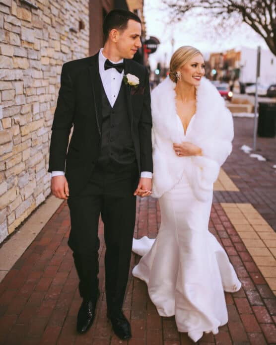 A bride in a white gown and fur shawl holds hands with a groom in a black tuxedo as they walk together on a brick sidewalk, smiling and looking at each other. by Justin Salem Meyer