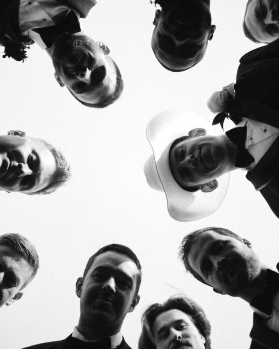 A group of men in formal wear, including one with a cowboy hat, stand in a circle and look down at the camera, photographed from below against a bright sky. The image is in black and white. by Justin Salem Meyer