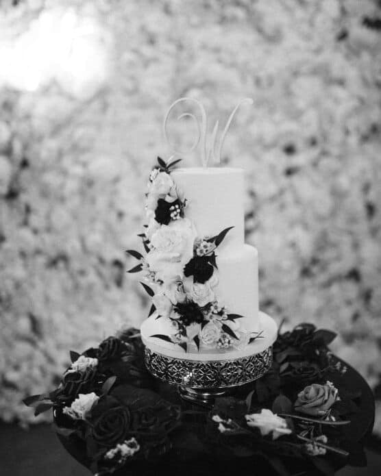 A two-tiered white cake decorated with white and dark flowers stands on an ornate stand. The cake features a cursive W topper and is surrounded by more flowers. The background is blurred with a floral texture. by Justin Salem Meyer