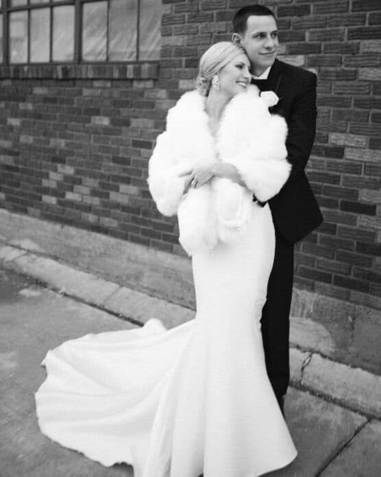 A bride in a white gown and fur shawl stands smiling with a groom in a dark suit, both embracing each other outside beside a brick building. by Justin Salem Meyer