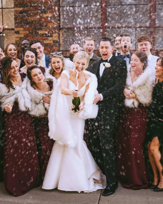 A bride and groom pop a champagne bottle, spraying bubbly as their joyful wedding party cheers behind them. Bridesmaids wear burgundy dresses with fur shawls, and everyone is smiling and celebrating outdoors. by Justin Salem Meyer