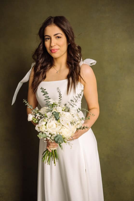 A woman in a white sleeveless dress holds a bouquet of white flowers and greenery. She has long, wavy brown hair and stands in front of a muted green background, looking at the camera with a slight smile. by Justin Salem Meyer