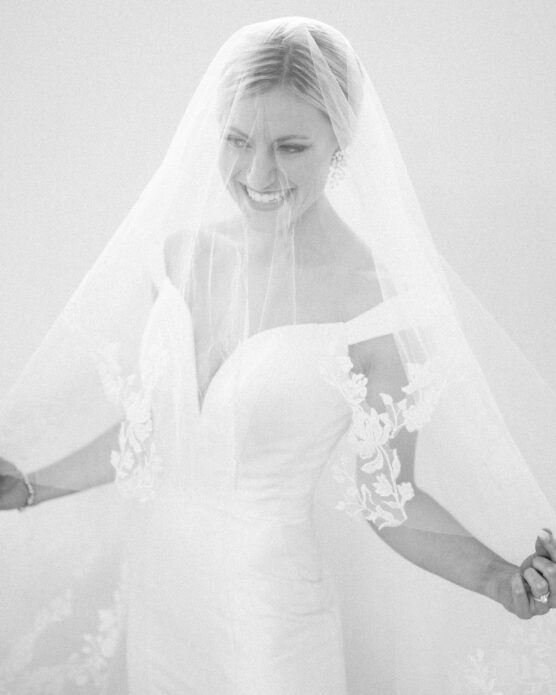 A smiling bride in an off-the-shoulder white wedding dress with floral lace details holds out her veil, which partially covers her face. The image is softly lit, creating an ethereal, elegant look. by Justin Salem Meyer