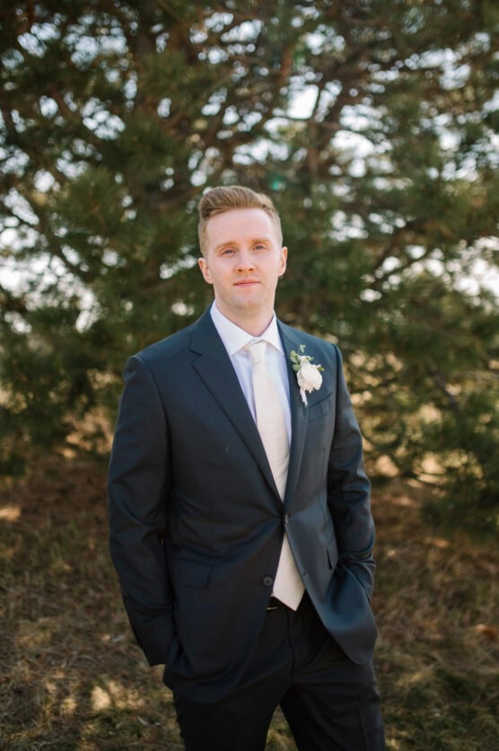 A man in a dark suit with a white shirt and tie stands outdoors in front of a tree, wearing a boutonnière and looking at the camera with his hands in his pockets. by Justin Salem Meyer