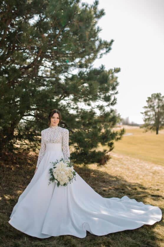A bride in a long-sleeved, high-neck white wedding dress stands outdoors by a tall pine tree, holding a bouquet of white flowers, with a grassy field and trees in the background. by Justin Salem Meyer