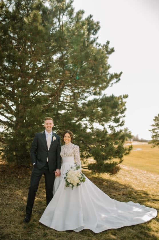 A bride in a long white gown holding a bouquet stands next to a groom in a dark suit. They are smiling and holding hands outdoors in front of a large pine tree on a sunny day. by Justin Salem Meyer