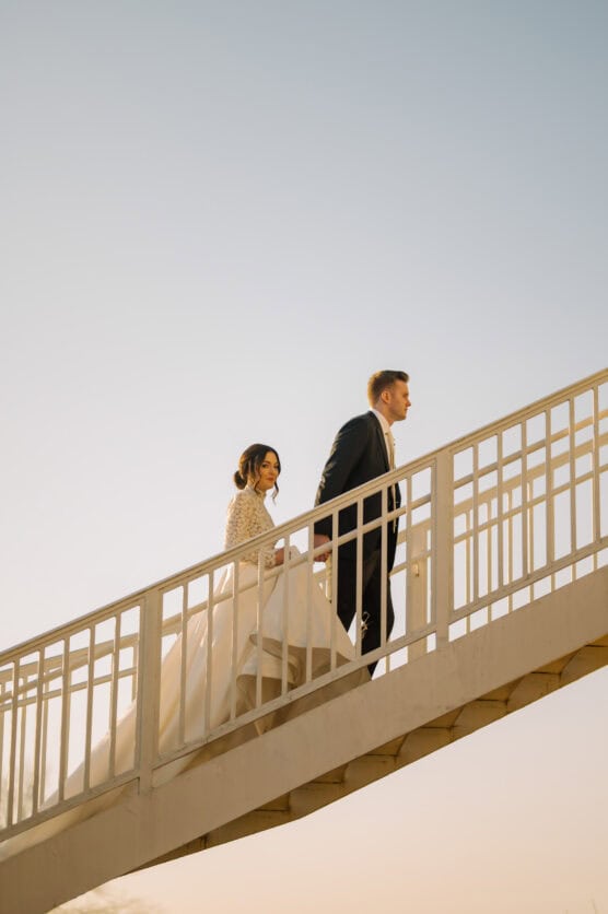 A bride and groom walk hand in hand up an outdoor staircase against a clear sky, with the bride wearing a white gown and the groom in a dark suit. by Justin Salem Meyer
