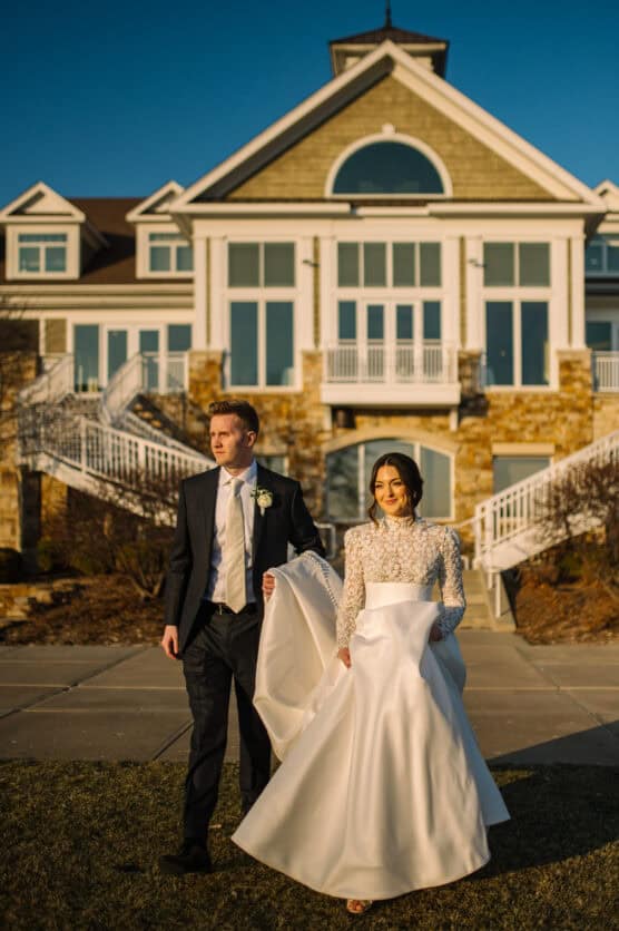 A bride in a long-sleeved white gown and a groom in a dark suit walk outside in front of a large, elegant house with big windows and stairs, illuminated by warm sunlight. by Justin Salem Meyer