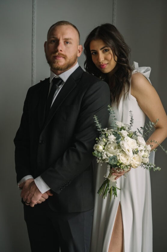 A man in a black suit stands in front of a woman in a white dress holding a bouquet of white flowers. They both face the camera with neutral expressions against a plain gray background. by Justin Salem Meyer