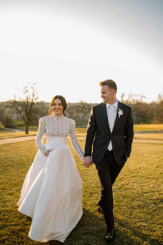 A bride in a long-sleeved, lace wedding dress and a groom in a black suit and tie walk hand in hand outdoors at sunset, smiling at each other on a grassy field. by Justin Salem Meyer