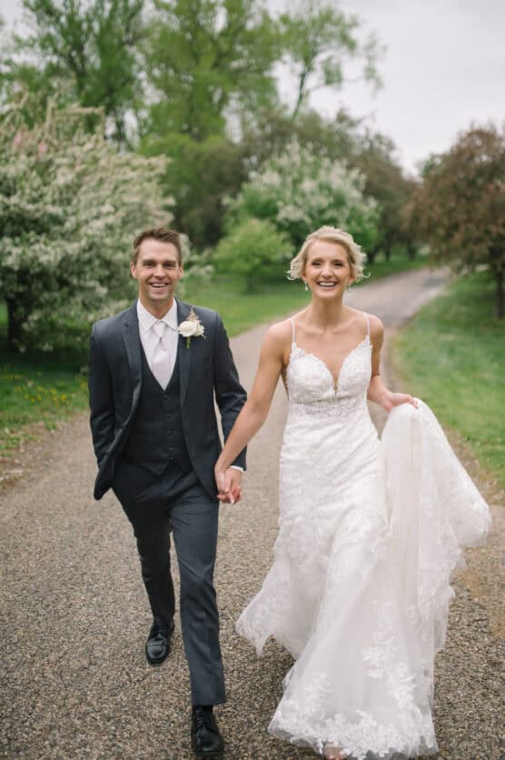 A joyful bride and groom walk hand in hand down a tree-lined path. The bride wears a white lace gown and holds her train, while the groom is in a dark suit with a white boutonniere. Both are smiling. by Justin Salem Meyer