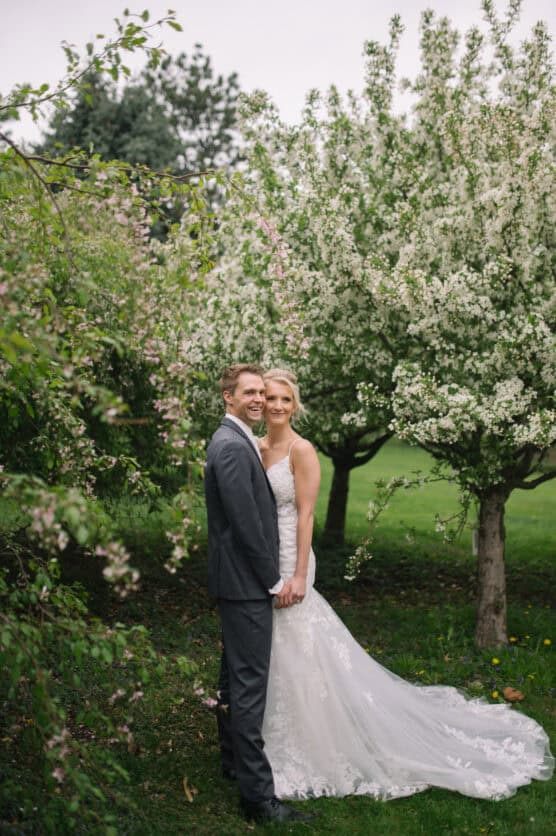 A bride and groom in wedding attire stand close together, smiling, surrounded by blooming trees and lush greenery in an outdoor garden setting. by Justin Salem Meyer