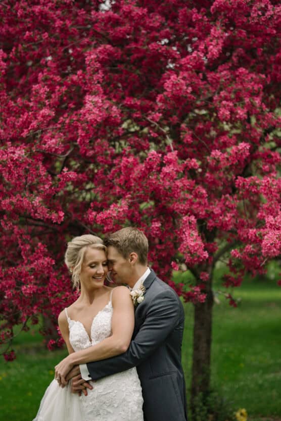 A bride and groom embrace and smile under a vibrant pink flowering tree, with lush greenery in the background. The bride wears a white lace dress, and the groom is in a gray suit. by Justin Salem Meyer