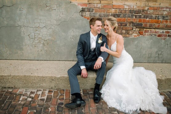 A bride in a white lace gown and a groom in a gray suit sit on a low concrete ledge, smiling and leaning close to each other, against a weathered brick and plaster wall. by Justin Salem Meyer