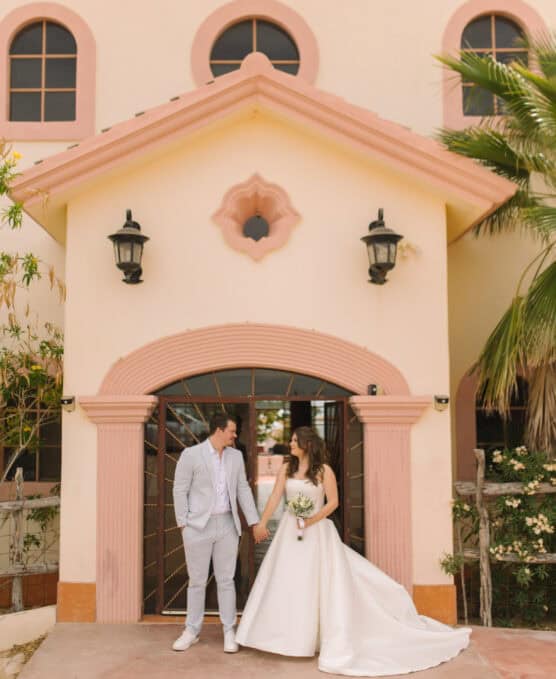 A bride in a white gown and a groom in a light suit stand holding hands and smiling at each other in front of a peach and cream building with arched windows and black lanterns. by Justin Salem Meyer