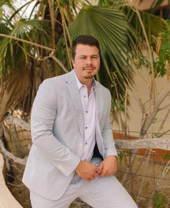 A man with short dark hair and a mustache, wearing a light gray suit and white shirt, leans casually against a wooden fence outdoors with palm leaves in the background. by Justin Salem Meyer
