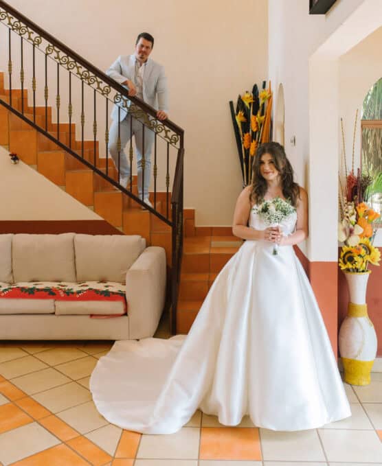 A bride in a white gown holds a bouquet and stands smiling indoors, while a groom in a light suit stands on a staircase behind her. The room has bright decor, tile flooring, and a beige sofa. by Justin Salem Meyer
