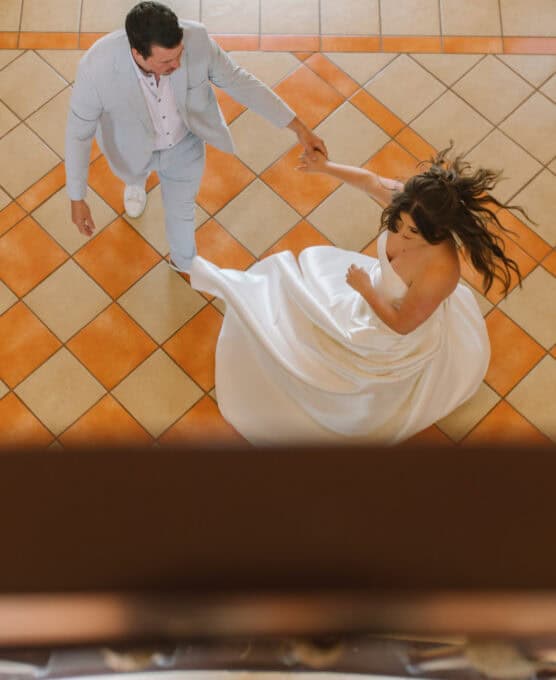 A bride in a white dress and a groom in a light suit dance together on a tiled floor, seen from above. The brides dress flows as they spin, and they hold hands joyfully. by Justin Salem Meyer