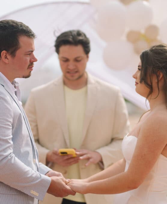 A bride and groom hold hands and face each other during their wedding ceremony, with an officiant standing between them holding a phone. White and gold balloons decorate the background. by Justin Salem Meyer