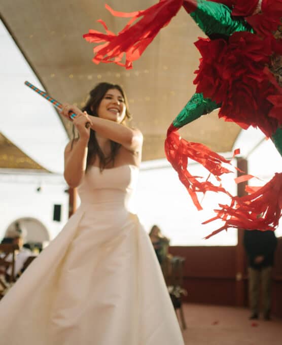 A smiling woman in a white dress swings a stick at a colorful piñata decorated with red and green tissue paper during an indoor celebration. by Justin Salem Meyer