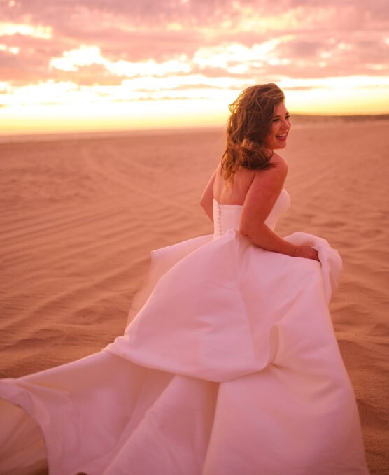 A woman in a flowing white wedding dress smiles as she looks back, walking across a sandy beach at sunset under a colorful sky. by Justin Salem Meyer
